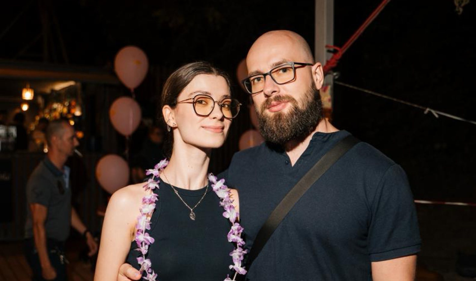 Max and a guest at an evening party, pink balloons and flower leis behind them