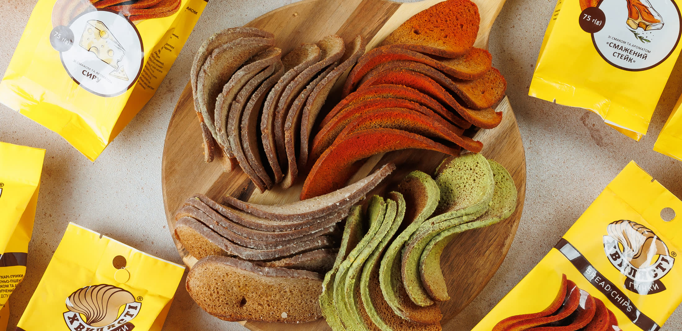 Wavy Khvyliasti bread chips fanned across a wooden board in four colours — rye, paprika red, pale wheat and herb green — ringed by the brand’s yellow retail packs