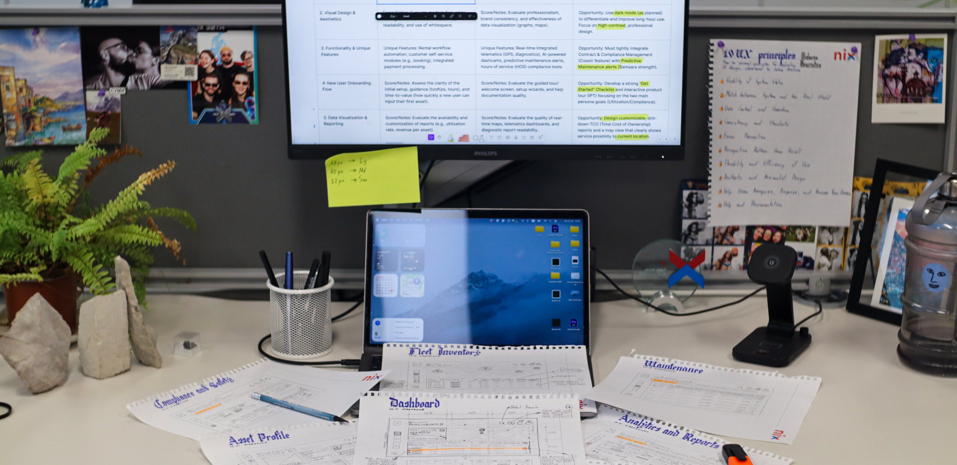 Max's desk mid-project — paper wireframes and notes spread below a laptop and a monitor showing a competitor-analysis board